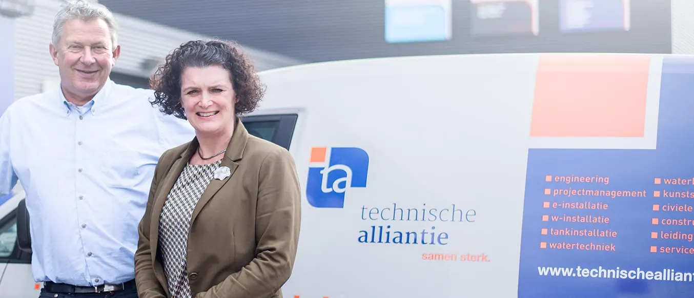 Twee medewerkers van Technische Alliantie bij de entree, naast een servicebus met het logo