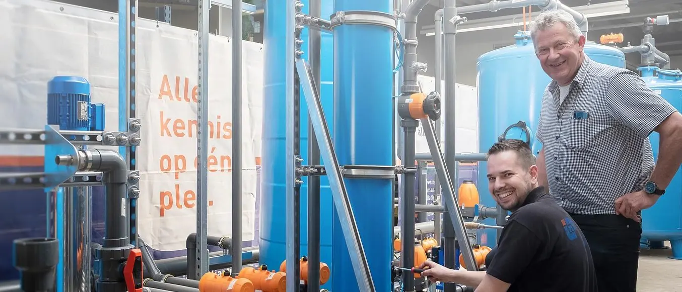 Twee medewerkers bij een waterbehandelingsinstallatie in opbouw in de werkplaats, met een spandoek op de achtergrond